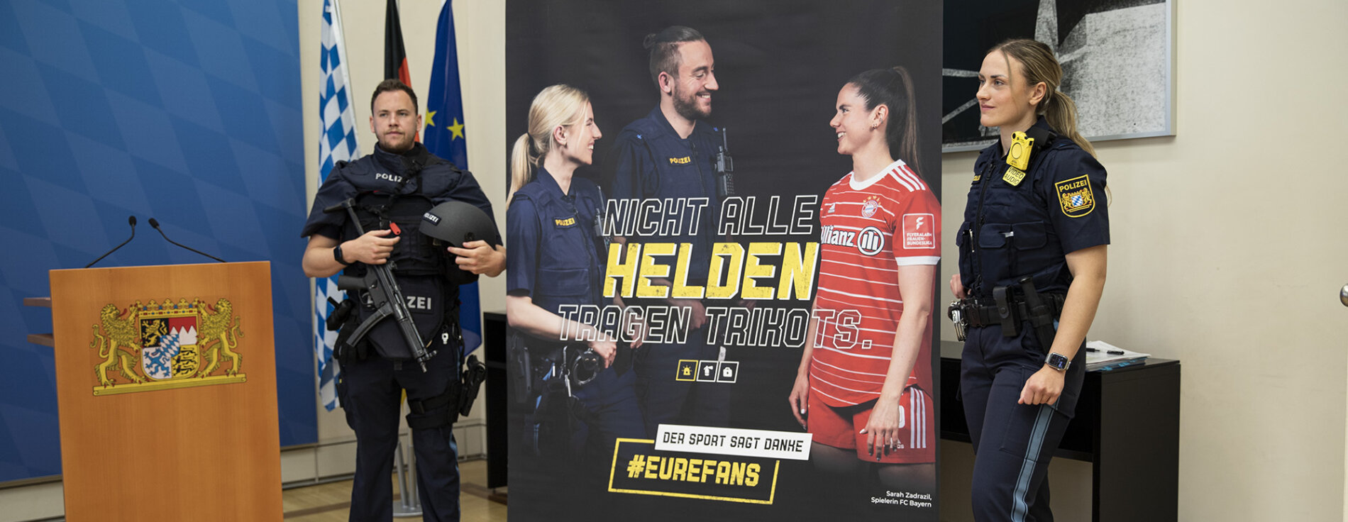 Polizistin und Polizist mit Schutzausrüstung neben Rollup zur Kampagne #EureFans für mehr Respekt für Einsatzkräfte